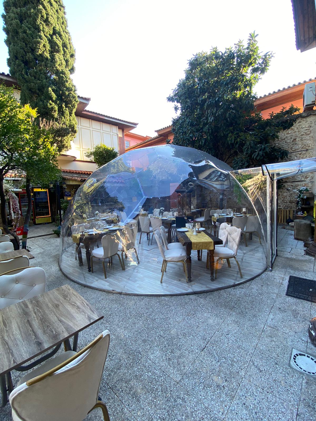Transparente Gartenkuppel mit offener Tür und gedeckten Tischen in einem begrünten Innenhof