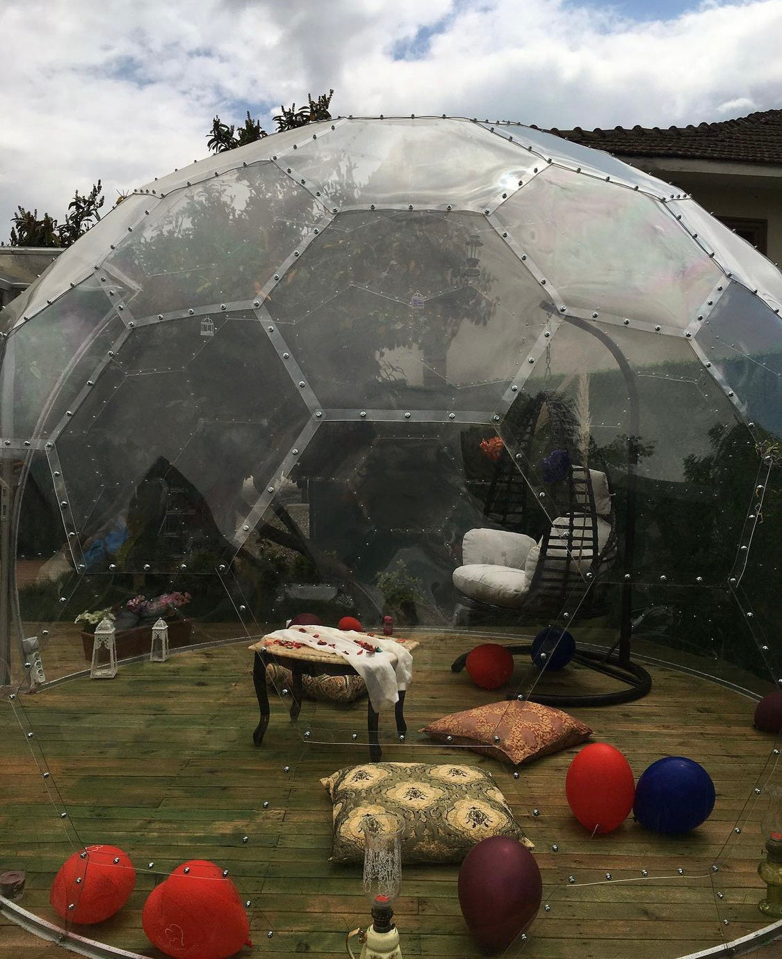Transparente Gartenkuppel auf einem Holzpodest mit Hängesessel, Kissen und Luftballons
